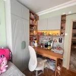 Bright study nook with a wooden desk, white chair, gray cabinets, and built‑in shelves filled with books and toys.