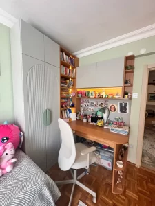 Bright study nook with a wooden desk, white chair, gray cabinets, and built‑in shelves filled with books and toys.