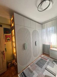 A contemporary gray wardrobe with large rounded doors and vertical grooves in a bright bedroom, with orange-lit shelves on the left.